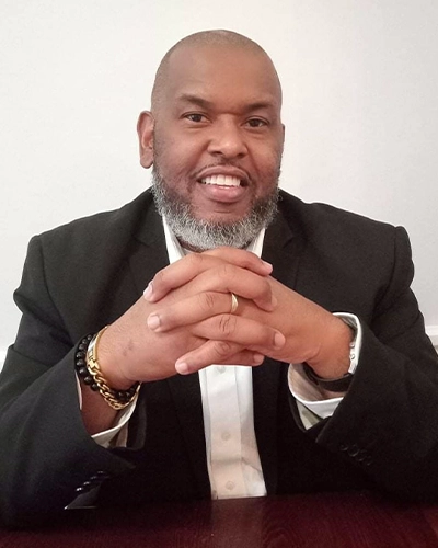 Chiropractor Torey Evans Smiling In A Suit With Hands Clasped At Desk